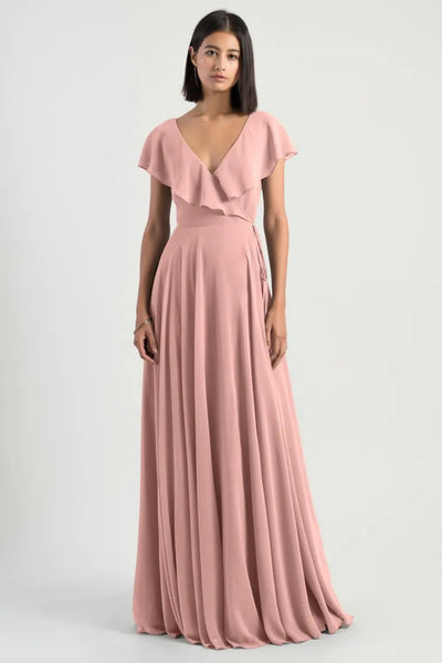 Woman in a romantic pink evening gown with flutter sleeves, the Faye - Bridesmaid Dress by Jenny Yoo from Bergamot Bridal, standing against a grey background.