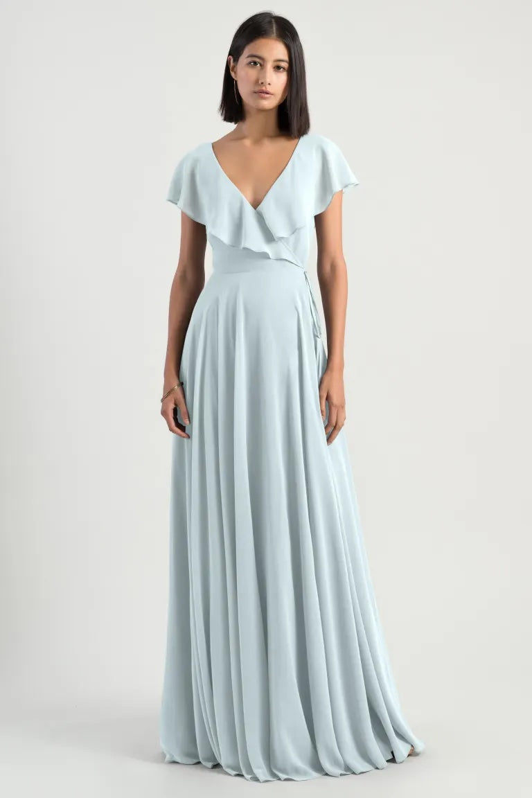 A woman modeling a pale blue, long formal Faye - Bridesmaid Dress by Jenny Yoo with a v-neckline and flutter sleeves from Bergamot Bridal.