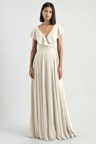 Woman in an elegant Faye - Bridesmaid Dress by Jenny Yoo chiffon wrap dress with ruffle flutter sleeves, standing against a neutral background from Bergamot Bridal.