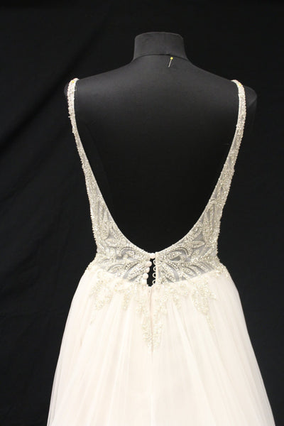 Back view of a mannequin displaying an elegant white V-neckline Justin Alexander Cady Dress - Off The Rack with detailed silver beadwork on the straps and waist, against a black background from Bergamot Bridal.