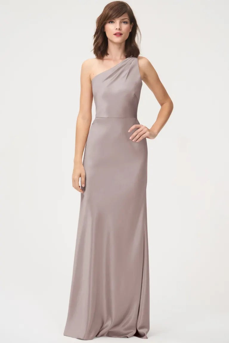 A woman wearing a satin back crepe, Lena - Bridesmaid Dress by Jenny Yoo with a one-shoulder neckline, floor-length in taupe from Bergamot Bridal.