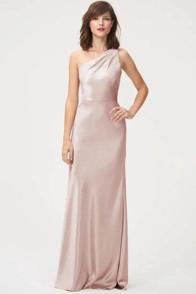A woman wearing a one-shoulder neckline, long blush-colored Bridesmaid Dress by Jenny Yoo stands posing for the camera.