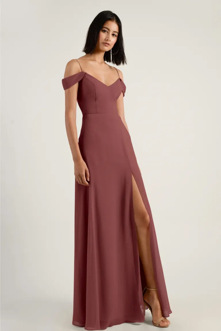 A woman in a stylish Priya - Bridesmaid Dress by Jenny Yoo with a side slit and off-shoulder sleeves stands against a neutral background from Bergamot Bridal.