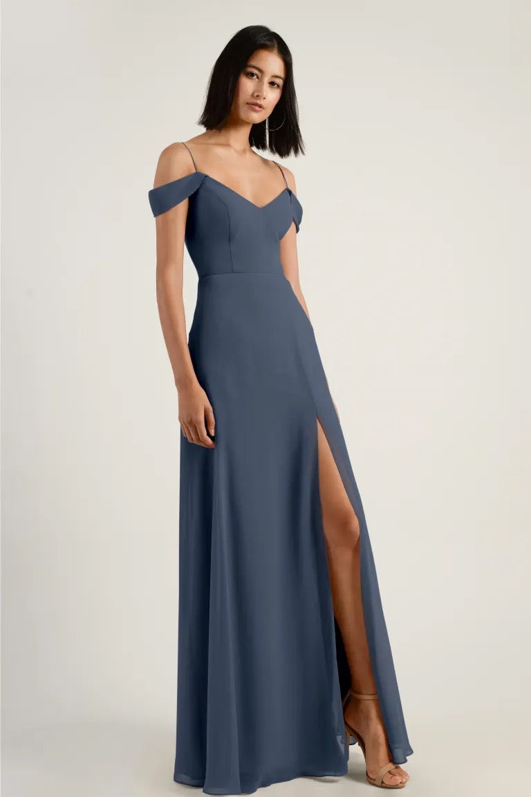 Woman in an elegant blue Priya V-neck evening gown by Jenny Yoo with an off-the-shoulder neckline and high leg slit from Bergamot Bridal.