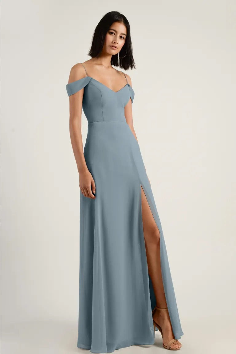 A woman modeling a light blue chiffon Priya bridesmaid dress by Jenny Yoo with a side slit and off-shoulder straps from Bergamot Bridal.
