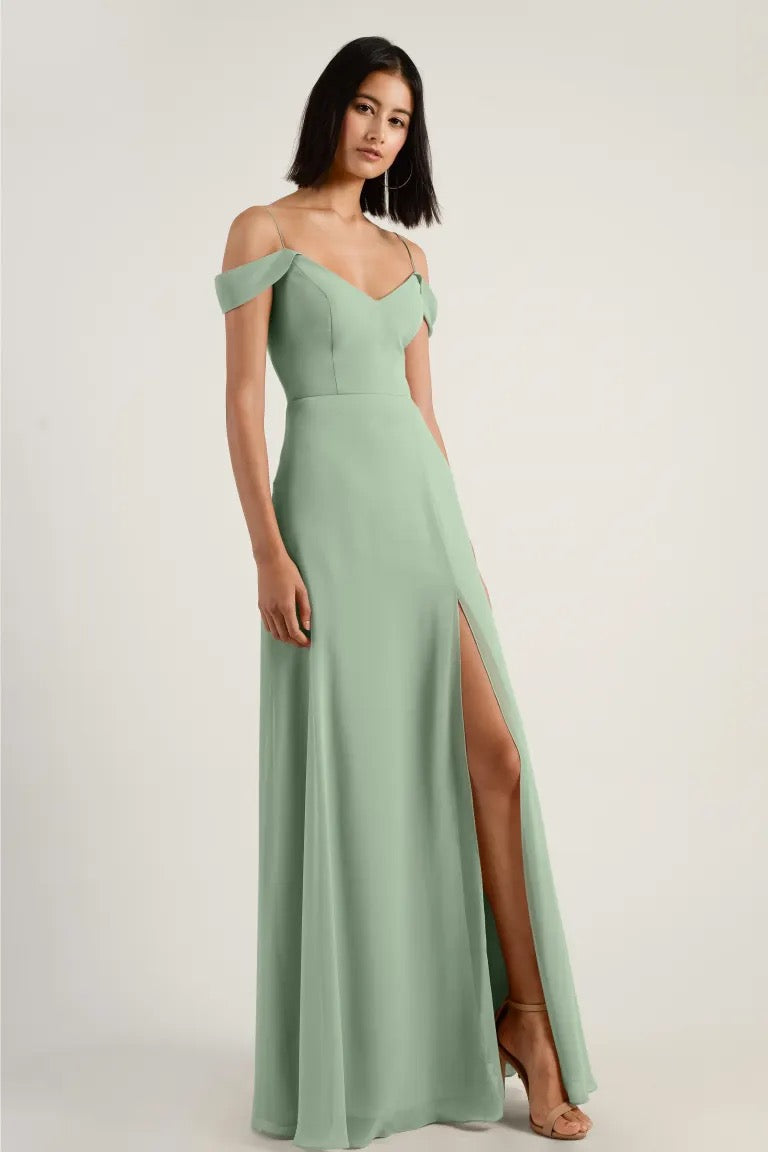 A woman in an elegant mint green chiffon off-the-shoulder bridesmaid dress with a high slit and A-line skirt, Priya - Bridesmaid Dress by Jenny Yoo from Bergamot Bridal.