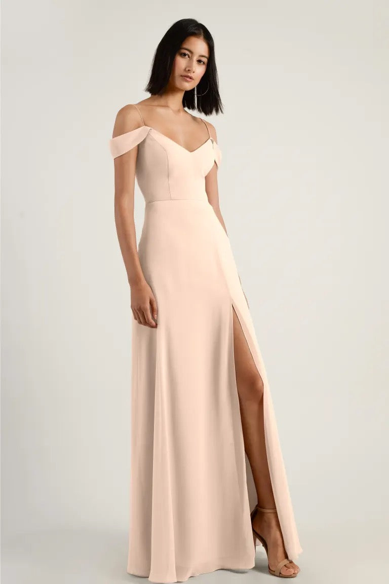 A woman models an elegant Priya - Bridesmaid Dress by Jenny Yoo with a high leg slit and off-the-shoulder sleeves from Bergamot Bridal.