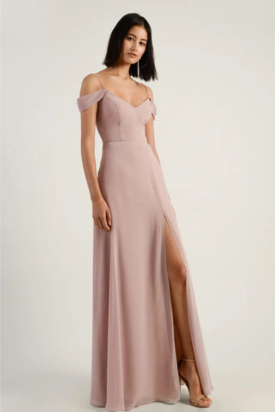 A woman in a pink Priya V-neck evening gown by Jenny Yoo with a thigh-high slit and off-the-shoulder sleeves poses against a neutral background.