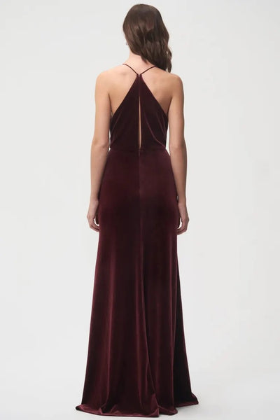 Woman standing facing away from the camera, wearing an elegant burgundy velvet bridesmaid dress with a halter cowl neckline and a floor-length hem by Bergamot Bridal.