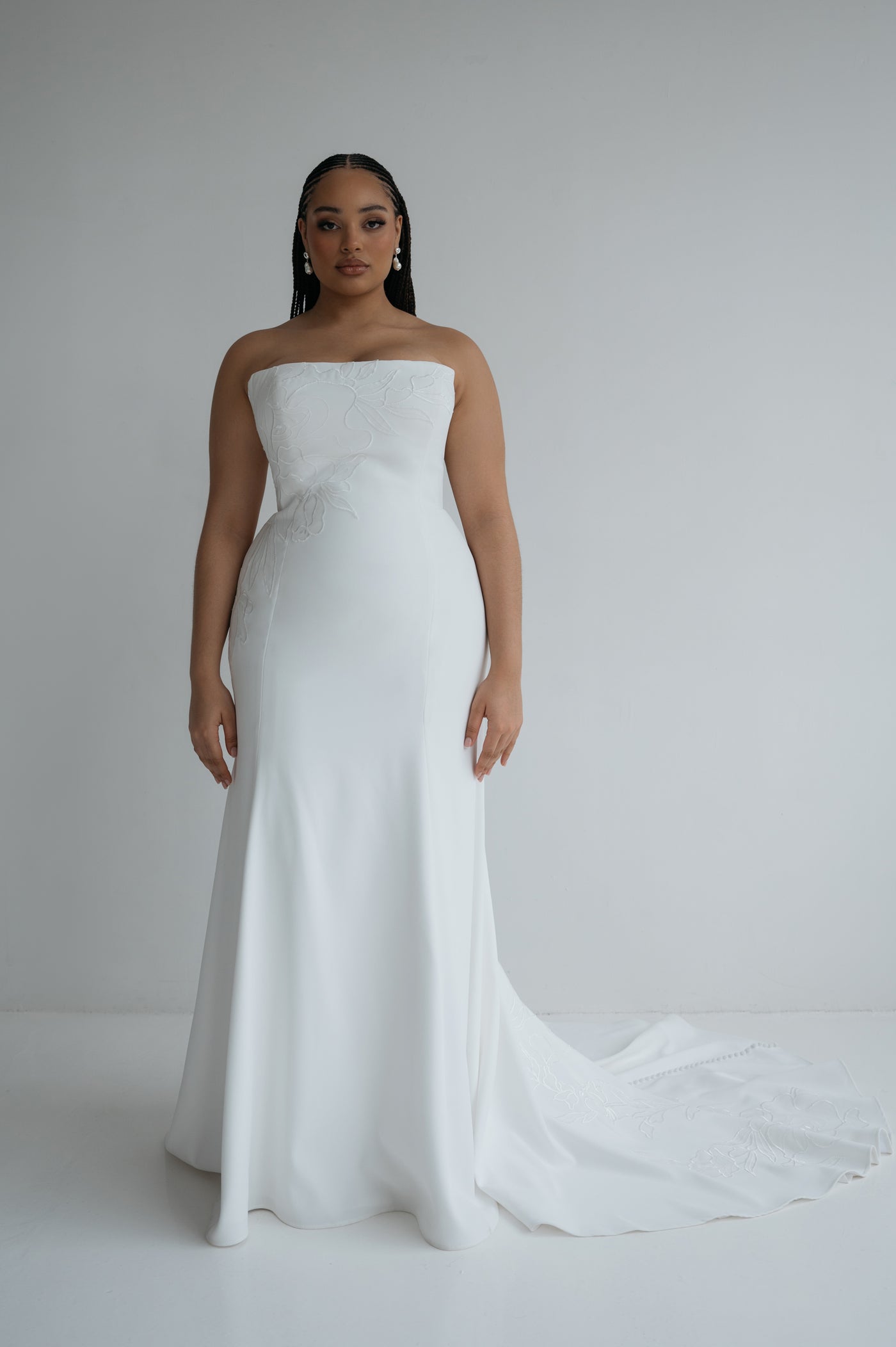 A woman poses in the Tricia Wedding Dress by Jenny Yoo for Bergamot Bridal, a strapless white gown with floral organza appliqué, fit and flare silhouette, and a slight train, set against a plain white background.