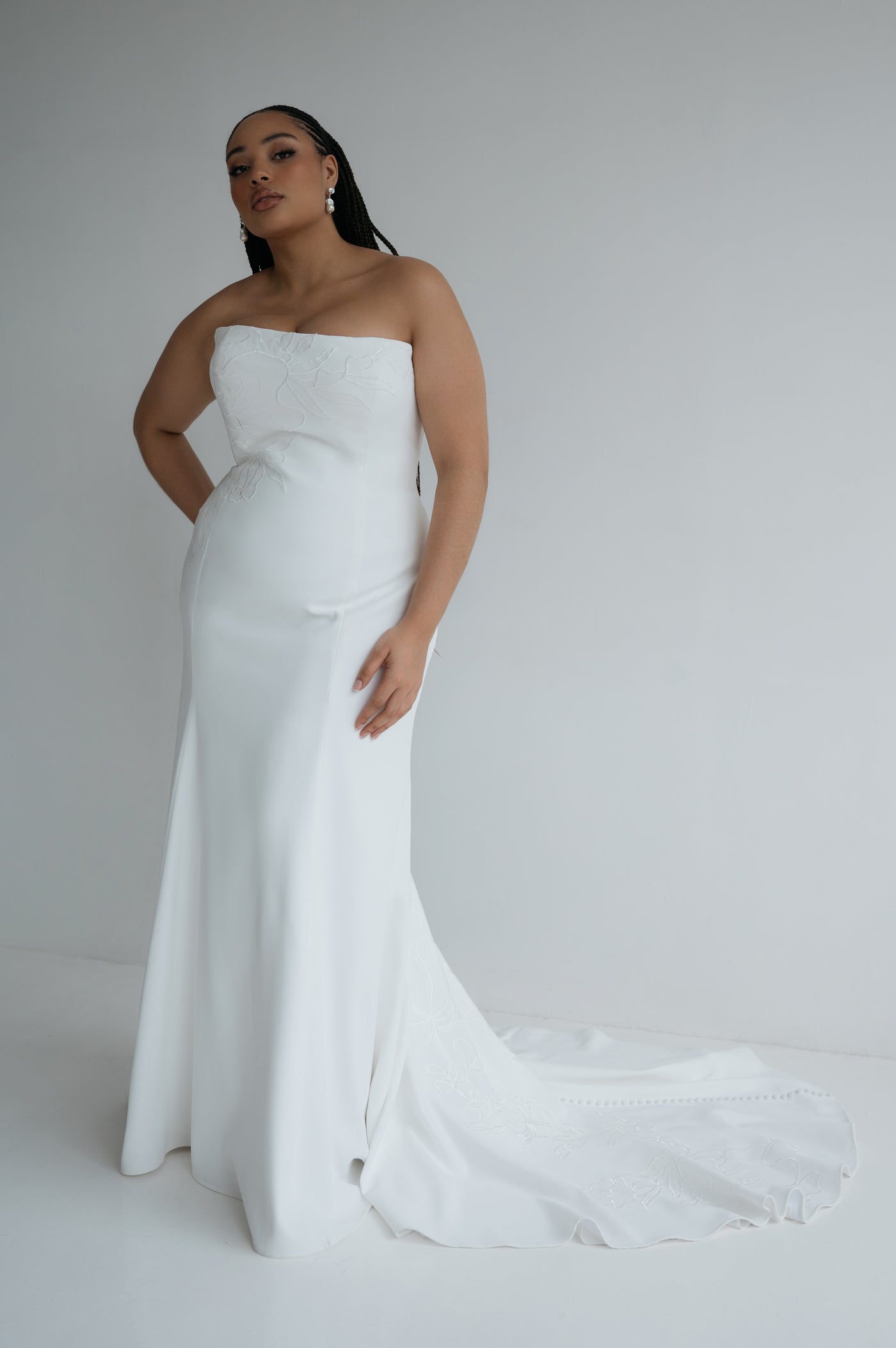 A woman poses against a plain light background wearing the Tricia Wedding Dress by Jenny Yoo for Bergamot Bridal, a strapless white gown featuring a fitted bodice, floral organza appliqué, and a slight train.