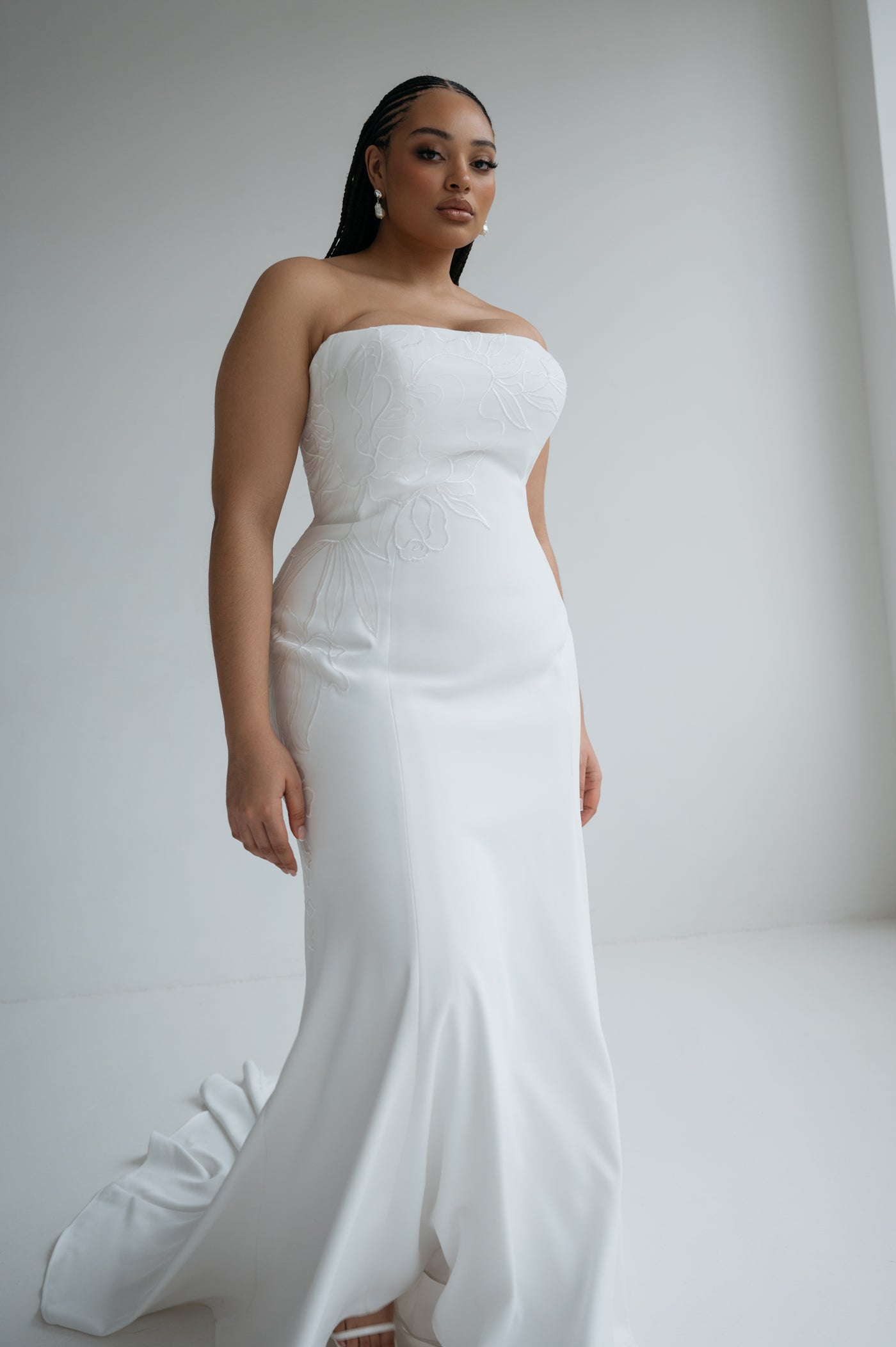 A woman wearing the Tricia Wedding Dress by Jenny Yoo from Bergamot Bridal—a strapless, fitted white gown with a fit and flare silhouette and delicate floral organza appliqué—stands in a minimalist, light-filled room.
