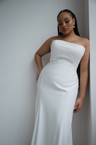 A woman in the Tricia Wedding Dress by Jenny Yoo for Bergamot Bridal, featuring a strapless fit and flare silhouette with floral organza appliqué, stands against a white wall, looking down with one hand at her side.
