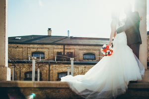 A bride and groom holding hands, silhouetted against a bright sky, with an industrial brick building in the background near bridal shops London.