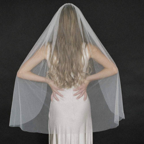 A woman in a white wedding dress and Ivory Plain Edge Bridal Veil by Bergamot Bridal stands with her back to the camera, hands on her lower back. Her long blond hair flows over the veil.