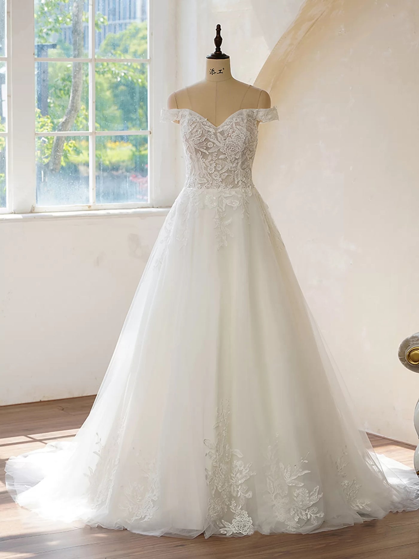 An Off the Shoulder Lace With Floral Wedding Dress by Bergamot Bridal in front of a window at a bridal shop.