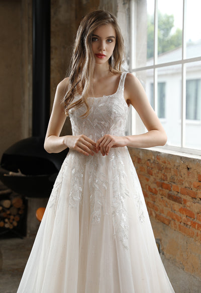 A woman in a white wedding dress, the Square Neckline Wedding Dress with Delicate Leafy Lace from Bergamot Bridal, is standing in front of a window at a bridal shop in London.