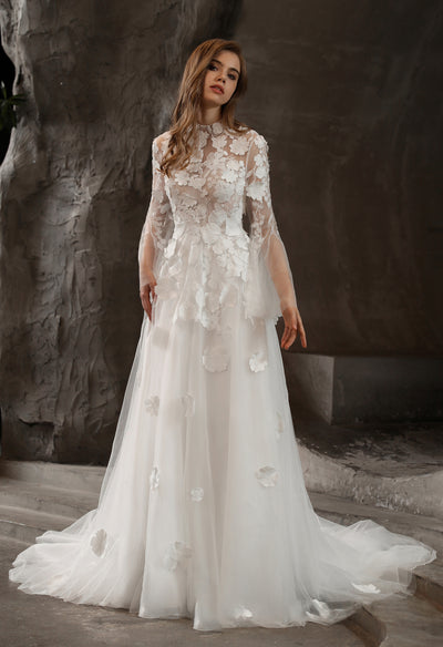 A bride in a white wedding dress, the Illusion High Neck Bridal Gown with Lovely 3D Floral Lace from Bergamot Bridal, is standing in front of a rock.