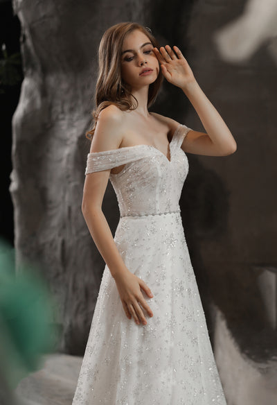 A woman in a white wedding dress, the Sparkly Beaded A-Line Bridal Gown With Off the Shoulder Sweetheart Neckline by Bergamot Bridal, is posing in front of a stone wall.