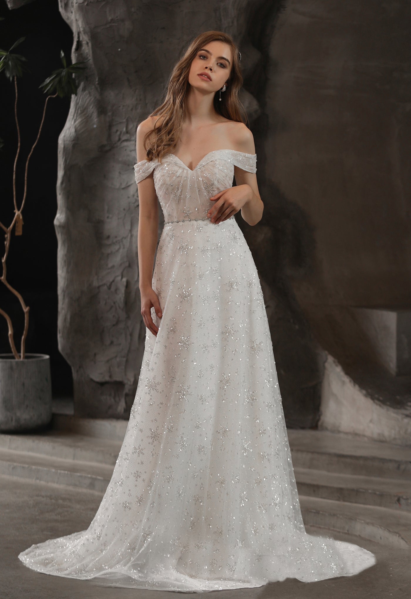 A woman in a white wedding dress from Bergamot Bridal, the Sparkly Beaded A-Line Bridal Gown With Off the Shoulder Sweetheart Neckline, is standing in front of a stone wall.