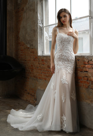 A woman in a Bergamot Bridal Square Neckline with Lace Straps Mermaid Wedding Gown - Off The Rack stands by a large window set against rustic brick walls, gazing outside.