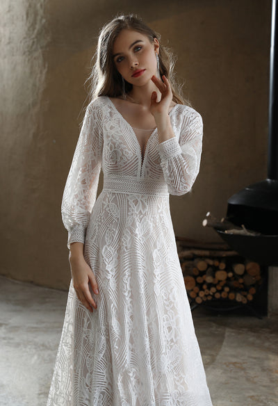 A woman in a Bergamot Bridal Plunging V-neck Lace Long Sleeve Bohemian Wedding Gown is posing in front of a fireplace at a bridal shop in London.