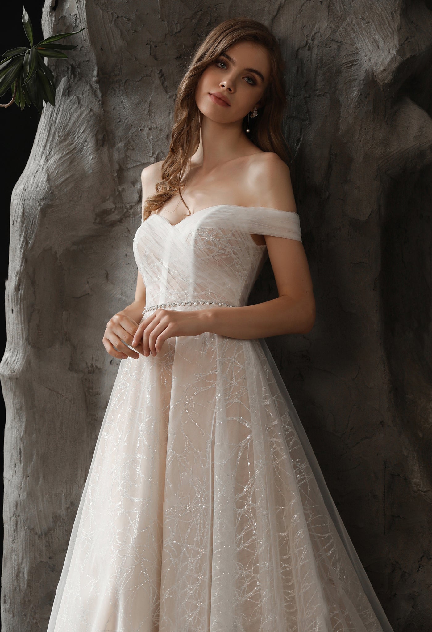 A woman in a Shimmer Off-the-shoulder Neckline A-line Tulle Wedding Dress by Bergamot Bridal posing in front of a stone wall at a bridal shop.