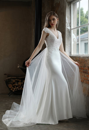 A woman wearing the Crepe Sheath Wedding Dress with Lace Cap Sleeves and Detachable Train from Bergamot Bridal stands near a window in a room adorned with rustic brick and wood accents.