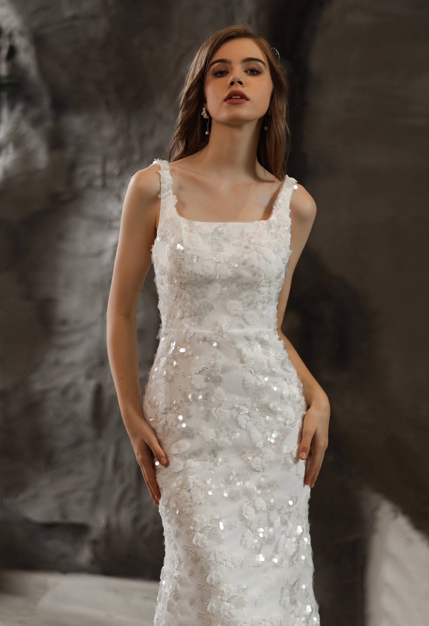 A woman in an elegant Bergamot Bridal wedding dress featuring sequined lace appliqués, posing against a textured grey background.