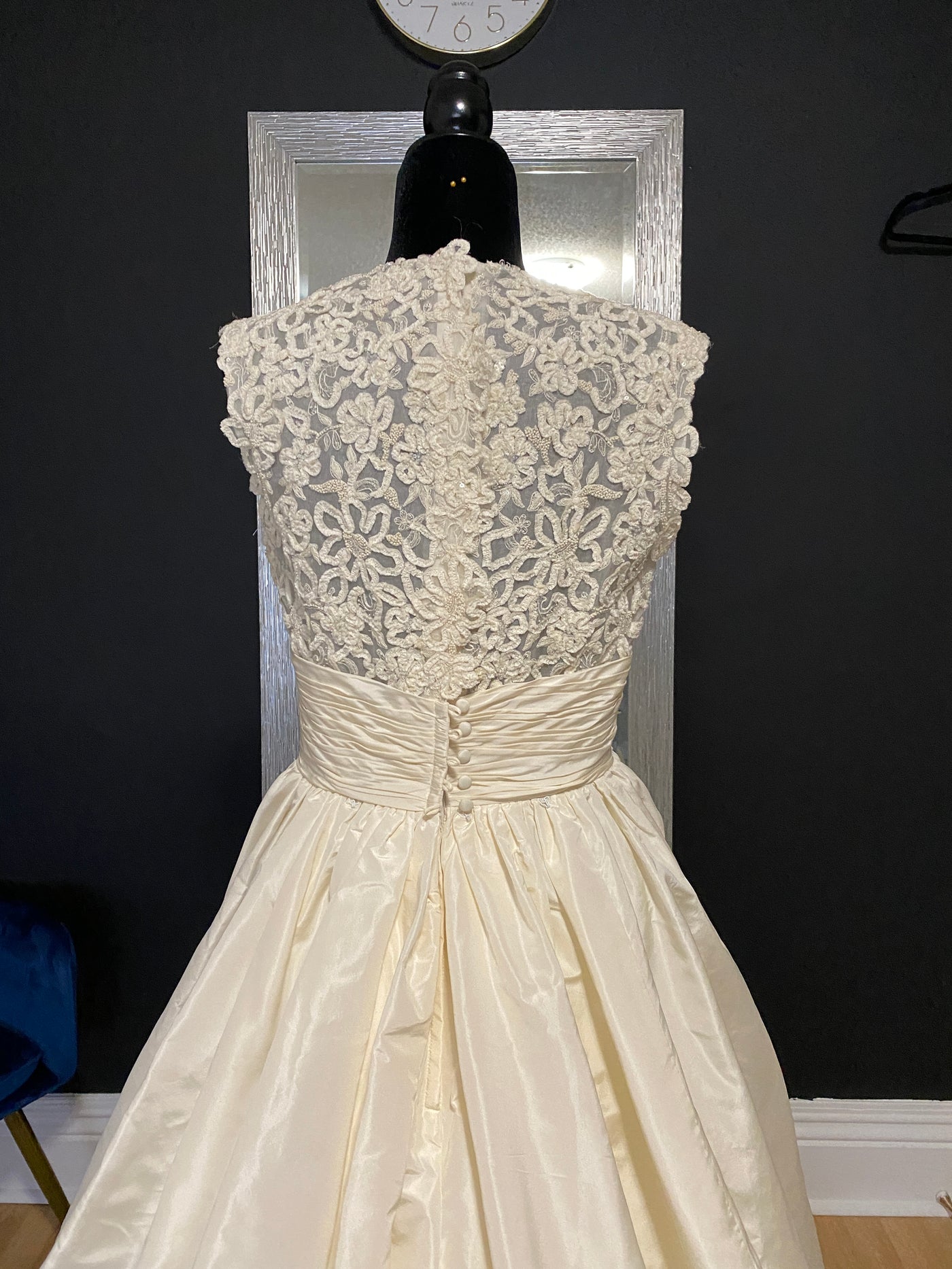 An elegant ivory Bergamot Bridal "Escalante" bridal gown with detailed lace bodice and gathered satin skirt on a mannequin against a dark wall with a clock above.