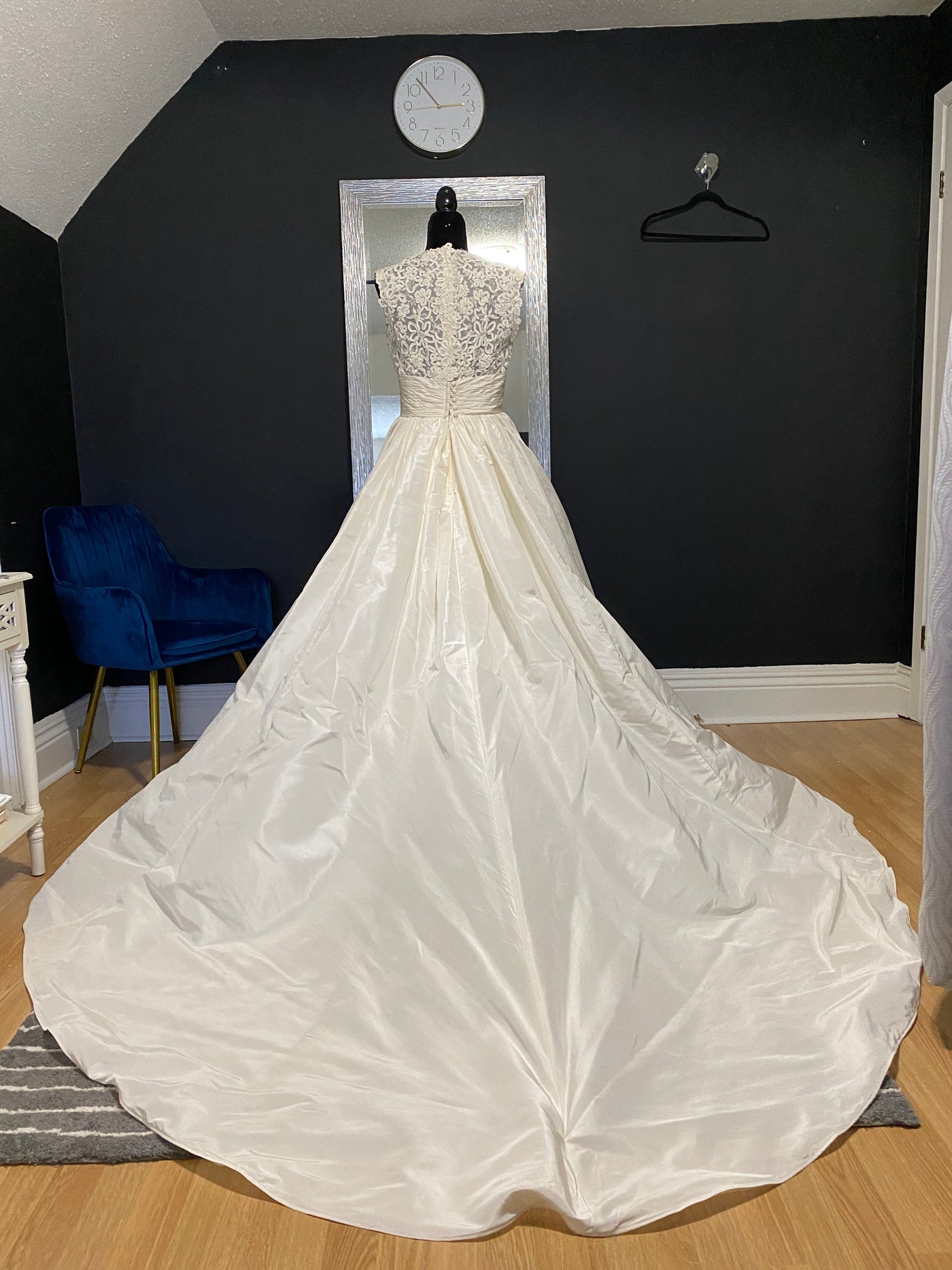 An elegant white Watters "Escalante" bridal gown with lace top and a cathedral train displayed in a room with dark walls and a blue chair.