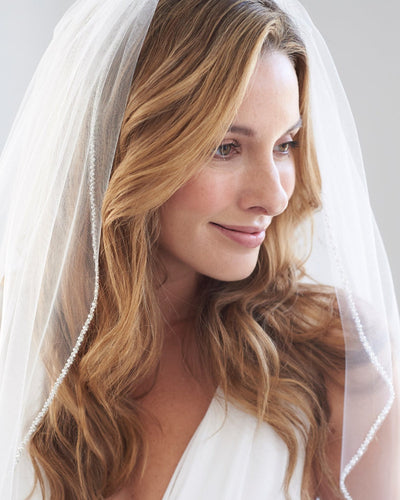 A bride with long, wavy hair under a Crystal Organza Beaded Wedding Veil from Bergamot Bridal, smiling gently in an off-white wedding dress, against a light background.