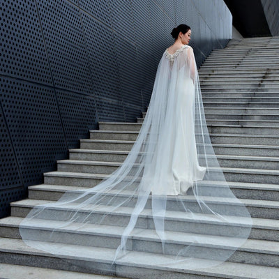 A bride in a flowing white wedding dress with a long veil stands on concrete stairs, with a modern, textured wall in the background wearing the Bergamot Bridal Bridal Cathedral Length Cape with Beaded Detail.