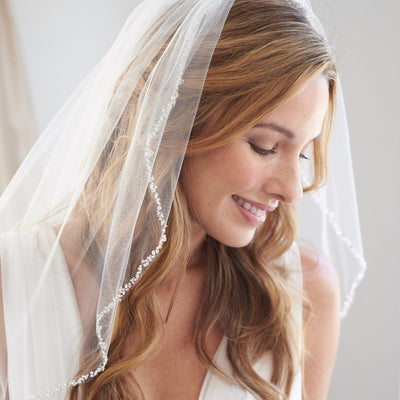 A smiling bride wearing a Bergamot Bridal Fingertip Length Crystal Beaded Veil, looking down gently in front of a soft, light background at one of the bridal shops in London Ontario.