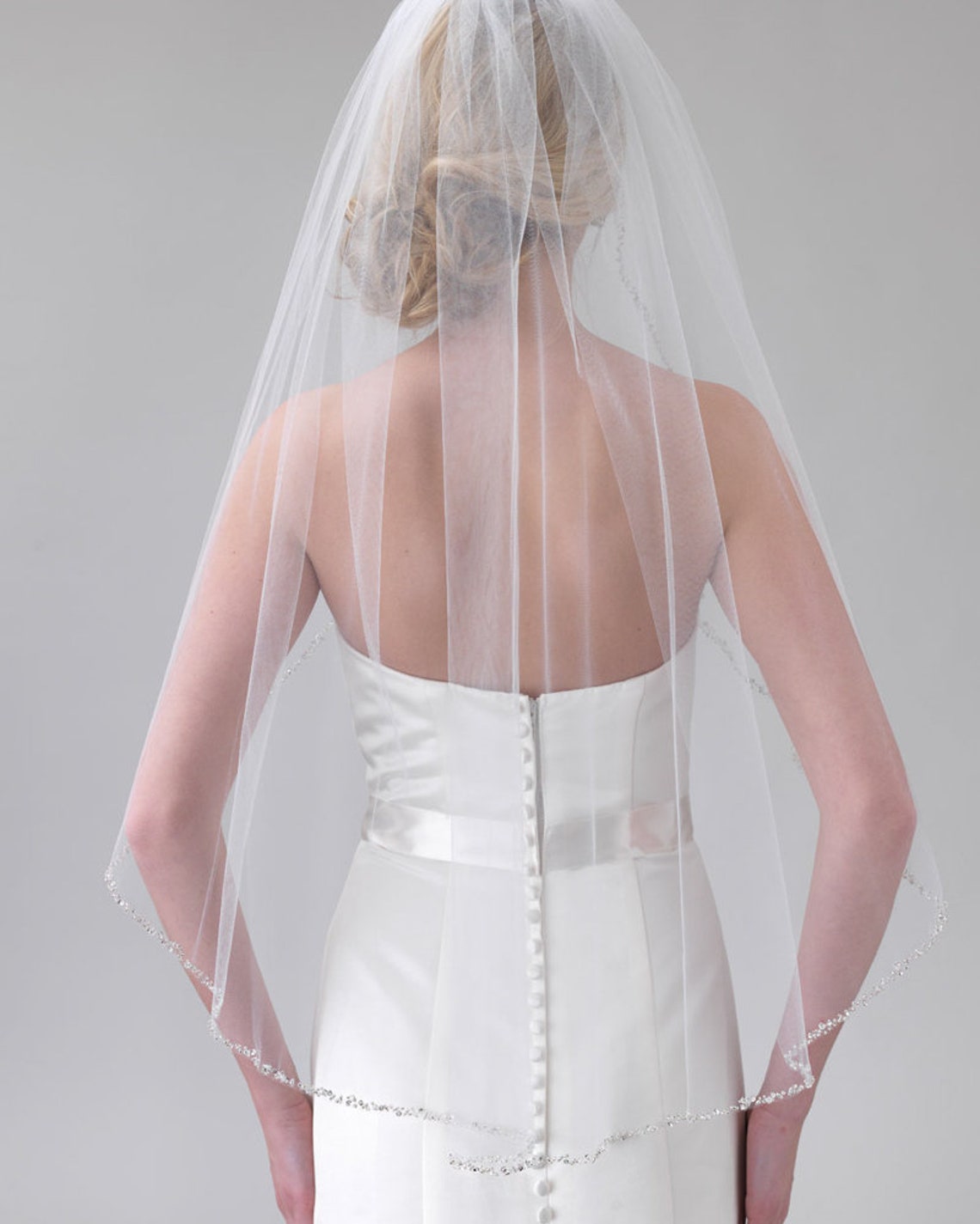 Back view of a bride wearing a white wedding dress from Bergamot Bridal and a Fingertip Length Crystal Beaded Veil, focusing on the detailed button closure of the dress.