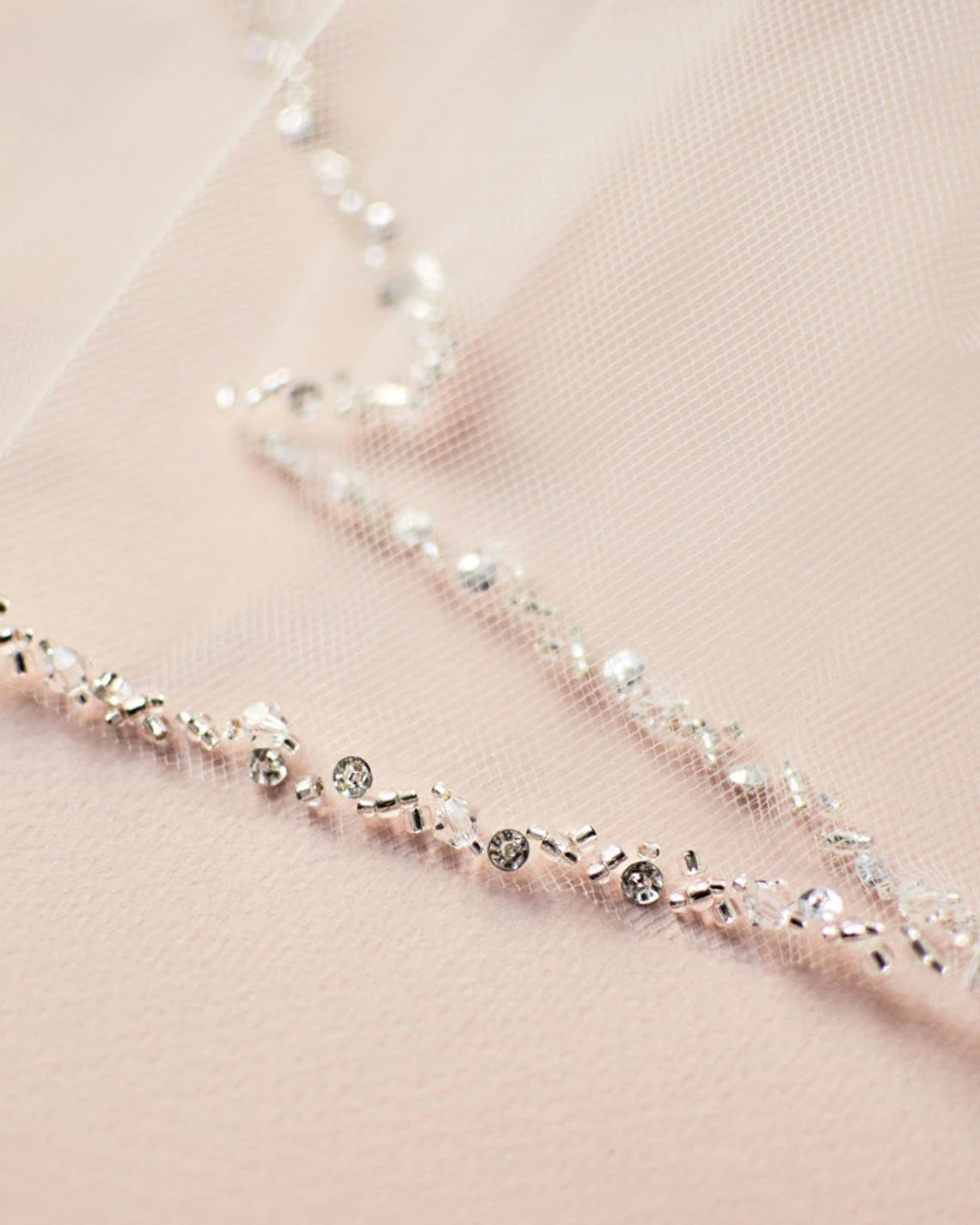Close-up of a delicate tulle fabric with intricate bead and sequin embellishments, perfect for bridal shops, on a soft pink background featuring the Bergamot Bridal Fingertip Length Crystal Beaded Veil.